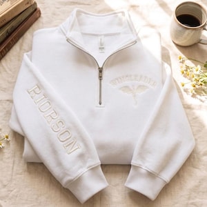 May include: A white quarter-zip sweatshirt featuring the embroidered text RIORSON vertically on the right sleeve and the text WINGLEADER with a dragon graphic embroidered in white on the upper left chest area near the metal zipper.