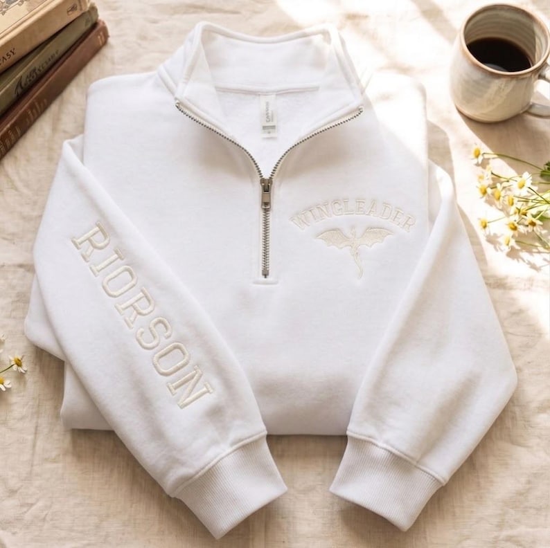 May include: A white quarter-zip sweatshirt featuring the embroidered text RIORSON vertically on the right sleeve and the text WINGLEADER with a dragon graphic embroidered in white on the upper left chest area near the metal zipper.