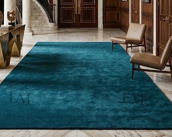 May include: A large rectangular teal area rug centered on a white marble floor in a grand foyer. The space features dark wood paneled walls, a tiered crystal chandelier, two tan chairs, a gold geometric console table, and marble columns.