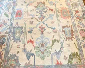 May include: A large, rectangular area rug with an intricate, traditional design. The rug features a cream-colored background with a border of blue and gray. The central pattern includes floral and geometric elements in shades of red, green, and brown.