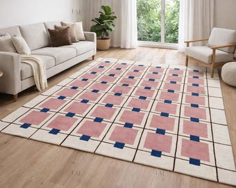 Op de afbeelding: Een rechthoekig vloerkleed met een geometrisch patroon. Het heeft een crèmekleurige achtergrond met een raster van zwarte lijnen. Binnen het raster bevinden zich roze vierkanten en blauwe rechthoeken. Het kleed ligt in een woonkamer.