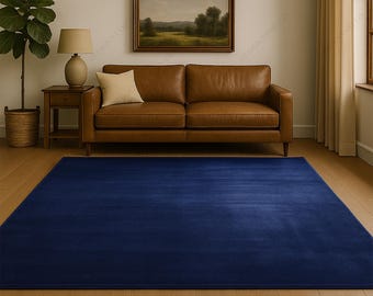 May include: A large, rectangular, solid navy blue rug dominates the foreground of a living room scene. A brown leather sofa sits against a cream-colored wall, with a landscape painting above it. A wooden side table with a lamp and a potted plant complete the decor.