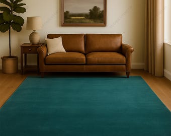 May include: A teal-colored rectangular rug in a living room setting. The rug is placed on a wooden floor, with a brown leather sofa, a side table with a lamp, and a framed landscape painting in the background. A large potted plant is visible.