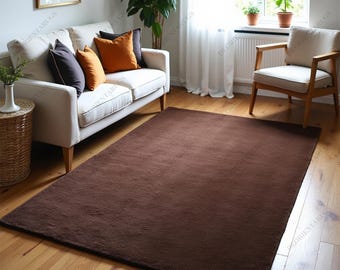 May include: A large, rectangular brown rug on a wooden floor. The rug is the centrepiece of a living room, with a white sofa, cushions, and a wooden chair in the background. Natural light enters the room through a window.
