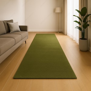 May include: A long, rectangular olive green rug on a hardwood floor. The rug is placed in front of a sofa and near a window with a tall plant. The room is well-lit with natural light.