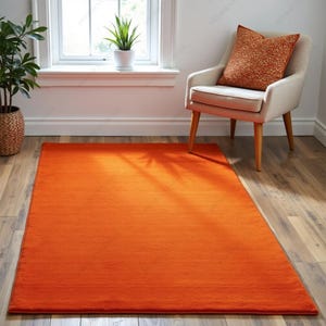 Puede incluir: Una alfombra rectangular naranja brillante se extiende sobre un suelo de madera. En el fondo, se observa un sillón crema con un cojín estampado. También se aprecia una ventana con una planta en maceta, creando un ambiente cálido.