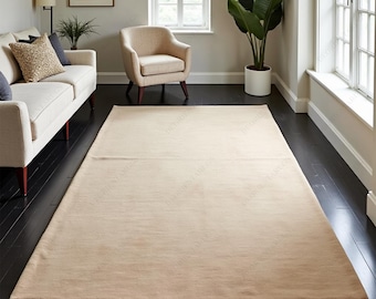 May include: A large, rectangular beige rug on a dark wood floor. The rug is positioned in front of a cream-colored sofa and a matching armchair. A large potted plant and a window provide natural light to the room.