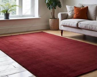 May include: A large, rectangular, deep red rug. The rug is positioned on a wooden floor in a living room, with a sofa, plants, and a window in the background. The rug's colour is a rich, solid shade of burgundy.