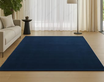 May include: A large, rectangular navy blue rug dominates the foreground of a living room. The rug is placed on a light-colored wooden floor, with a beige sofa and a black side table visible in the background. A floor lamp and sheer white curtains complete the scene.