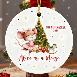 May include: A white ceramic Christmas ornament featuring a cartoon mouse in a pink tutu, with a Christmas tree and a rocking horse. Text reads "The Nutcracker 2024" and "Alice as a Mouse". Gold hanging cord.