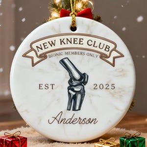 May include: A circular Christmas ornament with the words "NEW KNEE CLUB" and a knee joint graphic. It also features "BIONIC MEMBERS ONLY", "EST 2025", and the name "Anderson". Three small, wrapped gift boxes are at the base of the ornament, which is white with a marble effect.