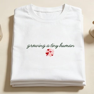 May include: White t-shirt featuring the text "growing a tiny human" in green script, with a cluster of red and pink hearts. The shirt is folded, with a small figurine and a diffuser visible in the background.