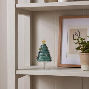 Op de afbeelding: Een decoratief kerstboombeeldje van glas in een doorschijnende blauwgroene kleur met een spiraalvormig ontwerp. De boom is voorzien van kleine, kleurrijke glazen kraalornamenten en wordt bekroond met een goudkleurige ster op een glazen voet.