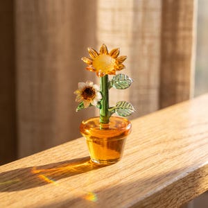 May include: A glass sculpture of a sunflower plant in a pot. The piece features a bright yellow sunflower bloom, a smaller white flower, and green glass leaves on a central stem. The entire structure is set in a translucent amber glass pot.