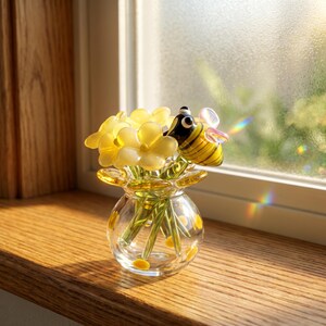 Puede incluir: Un florero de vidrio transparente que contiene un ramo de flores de vidrio amarillo y una pequeña figura de abeja de vidrio con rayas negras y amarillas y alas iridiscentes. El florero tiene una forma redondeada con puntos amarillos pintados.