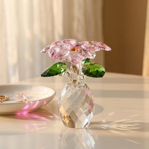 May include: A crystal figurine of a flower with light pink petals and a small amber center, featuring two deep green leaves. The flower is set in a clear, faceted, diamond-patterned crystal vase that reflects light across the surface.