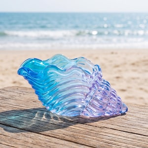 Peut inclure: Un bol décoratif en verre en forme de coquillage conque, présentant un dégradé translucide de teintes bleu vif et violet doux. La surface présente une texture ondulée et ridée qui imite les nervures naturelles d'un coquillage.