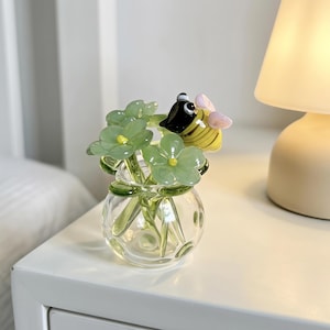 Puede incluir: Un pequeño florero de vidrio transparente que contiene varias flores de vidrio verde claro con centros amarillos y una figura de abeja de vidrio con rayas amarillas y negras, cabeza negra y alas rosa claro posada sobre el arreglo floral.