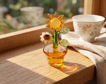 May include: A glass sculpture of two sunflowers in a small amber glass pot. The taller sunflower has orange petals and a textured centre, while the smaller one has white petals and a brown centre. A green glass stem features two green leaves.