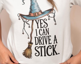 May include: White t-shirt featuring a witch hat and broom graphic. The hat is blue with a brown brim and buckle. The text on the shirt reads "YES I CAN DRIVE A STICK."