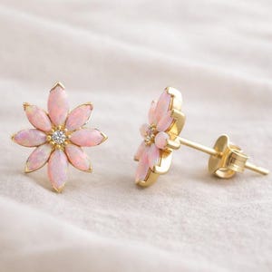 May include: A pair of gold-colored flower-shaped stud earrings. Each earring features pink petals surrounding a central clear gemstone. The earrings are set against a soft, off-white background.