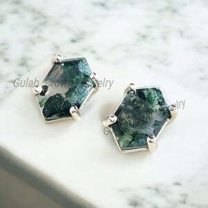 May include: A pair of hexagonal stud earrings. Each earring features a green moss agate stone set in a silver metal frame. The earrings are displayed on a white surface. The text "Gulab Crown Jewelry" is visible.