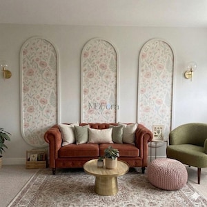 May include: A rust-colored tufted sofa with beige and sage green throw pillows, a round brass coffee table, a green armchair, and a pink knit pouf. Three arched wall panels feature a floral pattern with pink flowers and green foliage.