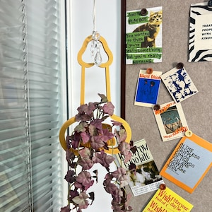 Puede incluir: Una maceta colgante de madera amarilla con un colgante de cristal, que contiene hiedra artificial púrpura en cascada. La maceta está suspendida por una cuerda blanca. Un tablón de anuncios con varias postales y notas está en el fondo.