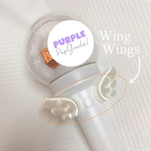 May include: A white light stick with a clear, glitter-filled globe top. The light stick has small, white wing-shaped attachments. The text "PURPLE PopGoods!" is in a circle on the top. The words "Wing Wings" are written in white.