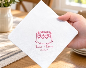 May include: A square white paper napkin featuring a pink line-art illustration of a tiered cake topped with cherries. Below the cake, the text reads Emma + Kevin followed by the date 11.02.27 in a matching pink script and print font.