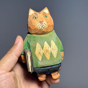 Może przedstawiać: Ręcznie rzeźbiona drewniana figurka kota w zielonym swetrze z ż&oacute;łtymi wzorami w romby. Kot ma pomarańczowe futro, zielone oczy i trzyma książkę. Unikalny, ręcznie robiony przedmiot dekoracyjny.