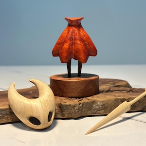 May include: A wooden sculpture set featuring a figure with a red cloak, a mask, and a small sword. The figure stands on a wooden base, and the mask and sword are placed nearby on a wooden surface. The set is crafted from light-coloured wood.