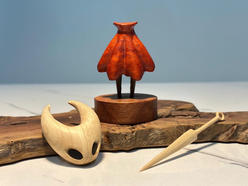 May include: A wooden sculpture set featuring a figure with a red cloak, a mask, and a small sword. The figure stands on a wooden base, and the mask and sword are placed nearby on a wooden surface. The set is crafted from light-coloured wood.