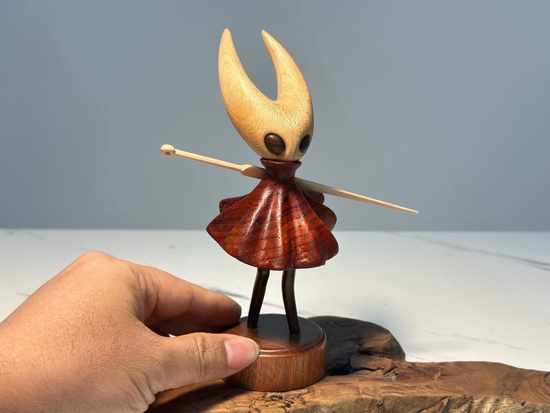 May include: A handcrafted wooden figurine of a character with a light-coloured head and horns, dark eyes, and a reddish-brown dress. The figure is mounted on a wooden base and has outstretched arms. The figurine is approximately 15 cm tall.