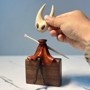 May include: A wooden figurine of a character with a light-coloured head and a dark red skirt, sitting on a brown block. The head is being held above the body by a hand. The figurine has a unique design.