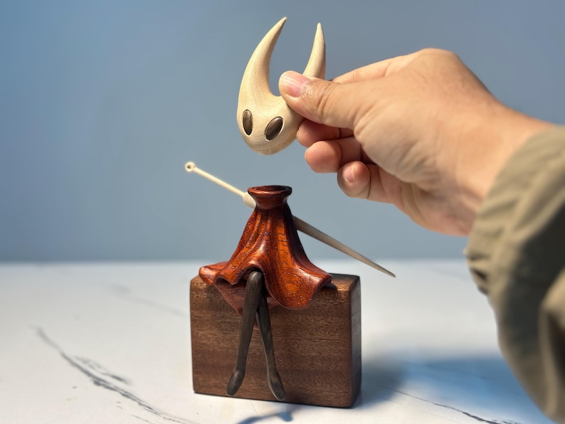 May include: A wooden figurine of a character with a light-coloured head and a dark red skirt, sitting on a brown block. The head is being held above the body by a hand. The figurine has a unique design.