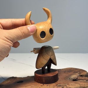 Peut inclure: Figurine en bois d'un personnage avec une t&ecirc;te &agrave; cornes et de grands yeux noirs. La figurine est faite de bois brun clair et fonc&eacute;, avec une base et une structure en forme de cape. La t&ecirc;te est tenue au-dessus du corps.