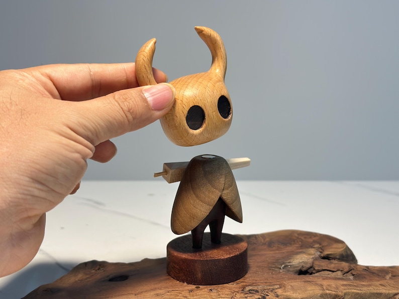 Peut inclure: Figurine en bois d'un personnage avec une t&ecirc;te &agrave; cornes et de grands yeux noirs. La figurine est faite de bois brun clair et fonc&eacute;, avec une base et une structure en forme de cape. La t&ecirc;te est tenue au-dessus du corps.