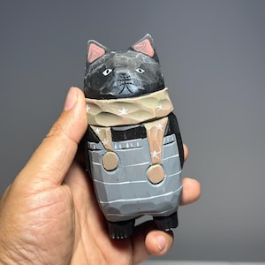 May include: A hand-carved wooden cat figurine. The black cat has pink-lined ears, white eyes, and a tan scarf with white stars. It wears gray overalls with tan buttons and white star accents. The figurine is held in a person's hand.