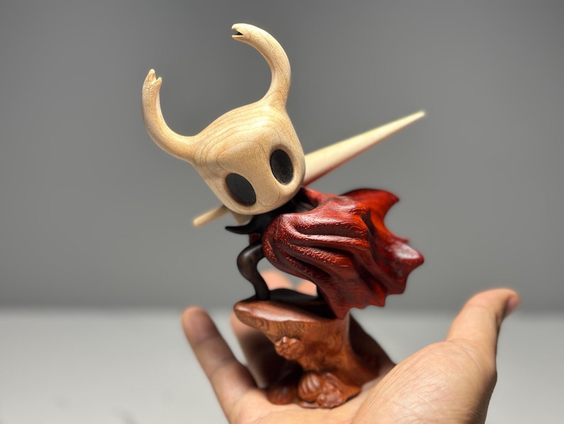 Peut inclure: Figurine en bois d'un personnage avec une t&ecirc;te de couleur claire, de grands yeux noirs et une cape rouge. La figurine tient une longue arme pointue. La figurine est sur une base en bois.
