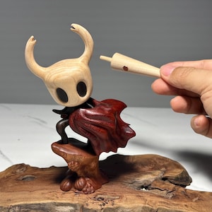 Peut inclure: Sculpture en bois d'un personnage avec une t&ecirc;te claire et une cape rouge fonc&eacute;. La figurine est mont&eacute;e sur une base en bois. Une main tient une pi&egrave;ce en bois pr&egrave;s de la t&ecirc;te du personnage. Il s'agit probablement d'un objet de collection ou d'une &oelig;uvre d'art.
