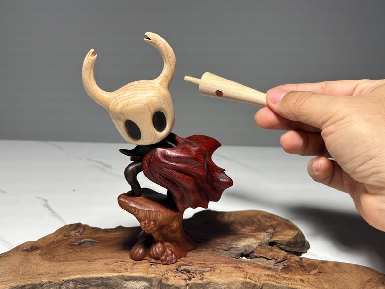Peut inclure: Sculpture en bois d'un personnage avec une t&ecirc;te claire et une cape rouge fonc&eacute;. La figurine est mont&eacute;e sur une base en bois. Une main tient une pi&egrave;ce en bois pr&egrave;s de la t&ecirc;te du personnage. Il s'agit probablement d'un objet de collection ou d'une &oelig;uvre d'art.