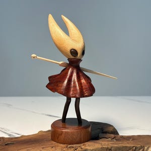 May include: A wooden figurine of a character with a light-coloured head and two pointed horns. The figure has a dark red dress and outstretched arms. It stands on a wooden base, showcasing detailed craftsmanship and a unique design.