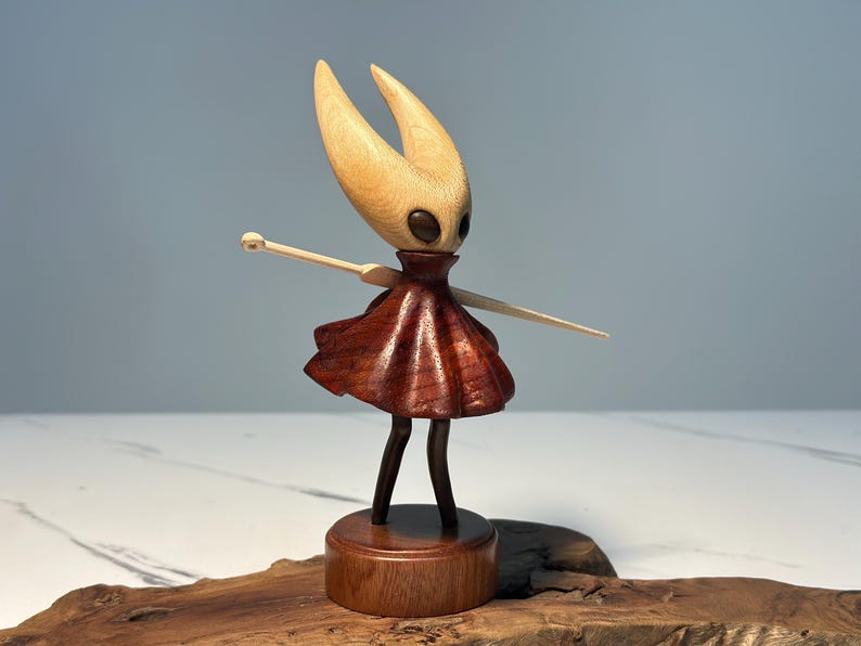 May include: A wooden figurine of a character with a light-coloured head and two pointed horns. The figure has a dark red dress and outstretched arms. It stands on a wooden base, showcasing detailed craftsmanship and a unique design.