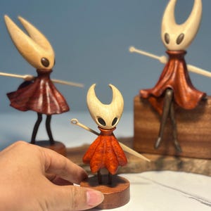 May include: Wooden figurines of a character with a light-coloured head, dark eyes, and a red dress. The figurines have outstretched arms and are on wooden bases. One figurine is held by a hand.
