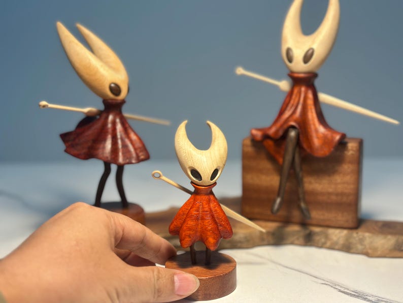 May include: Wooden figurines of a character with a light-coloured head, dark eyes, and a red dress. The figurines have outstretched arms and are on wooden bases. One figurine is held by a hand.