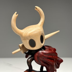 Peut inclure: Figurine en bois d'un personnage avec une t&ecirc;te en bois clair, de grands yeux noirs et des cornes incurv&eacute;es. Il porte une &eacute;p&eacute;e en bois et une cape rouge fonc&eacute;. Objet de collection.