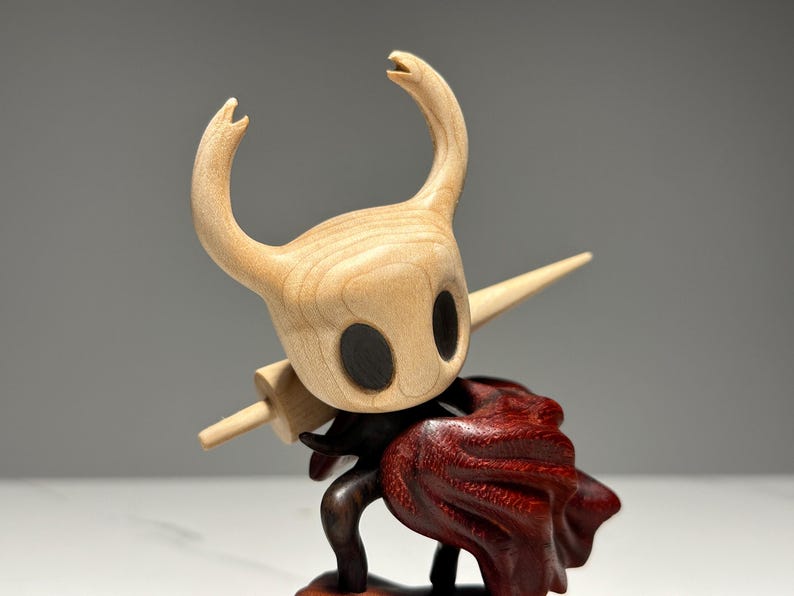 Peut inclure: Figurine en bois d'un personnage avec une t&ecirc;te en bois clair, de grands yeux noirs et des cornes incurv&eacute;es. Il porte une &eacute;p&eacute;e en bois et une cape rouge fonc&eacute;. Objet de collection.