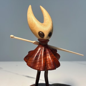 May include: Wooden figurine of a character with a light-coloured head and horns, dark eyes, and a reddish-brown dress. The figure has outstretched arms with a light-coloured wooden staff. The figurine is standing on a dark base.
