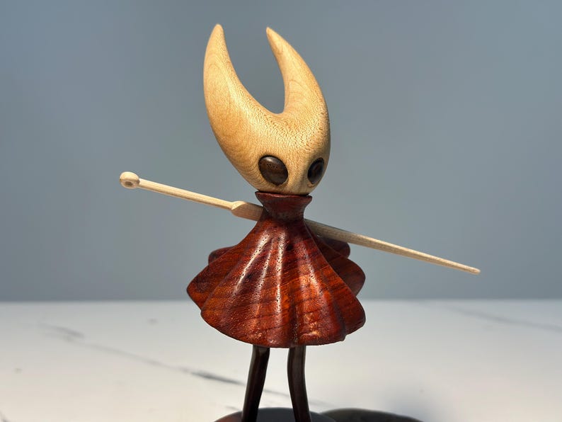 May include: Wooden figurine of a character with a light-coloured head and horns, dark eyes, and a reddish-brown dress. The figure has outstretched arms with a light-coloured wooden staff. The figurine is standing on a dark base.